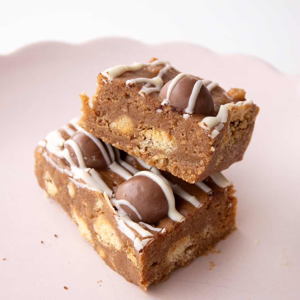 Two stacked square chocolate chip cookie bars with white drizzle and milk chocolate balls on a pale pink plate.