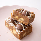 Two stacked square chocolate chip cookie bars with white drizzle and milk chocolate balls on a pale pink plate.