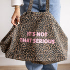 A person wearing blue jeans and a white top holds up a large leopard print tote bag with pink text that reads 'IT'S NOT THAT SERIOUS'.
