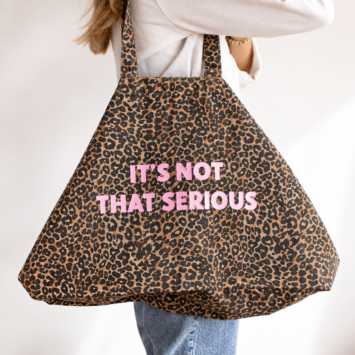 A person carrying a leopard print tote bag with the text 'IT'S NOT THAT SERIOUS' in pink lettering, shown against a plain white background.
