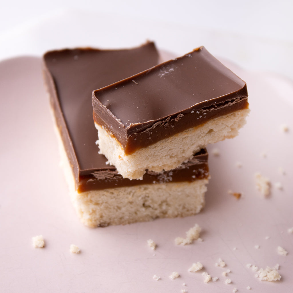 Two chocolate-topped cookie bars stacked on a pale pink plate, with crumbs scattered around on a light background.