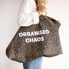 A person wearing a white sweatshirt and blue jeans carries a large leopard-print tote bag with the words 'ORGANISED CHAOS' printed in bold white letters on the front, against a plain light background.