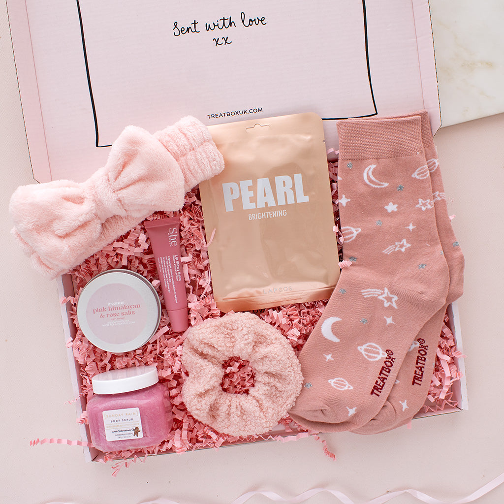 A curated self-care gift box filled with pink and peach-toned items, including a plush eye mask, scrunchie, socks, a tube, a jar, and a pouch labeled PEARL BREATHEING, arranged in pink shredded paper inside an open box.