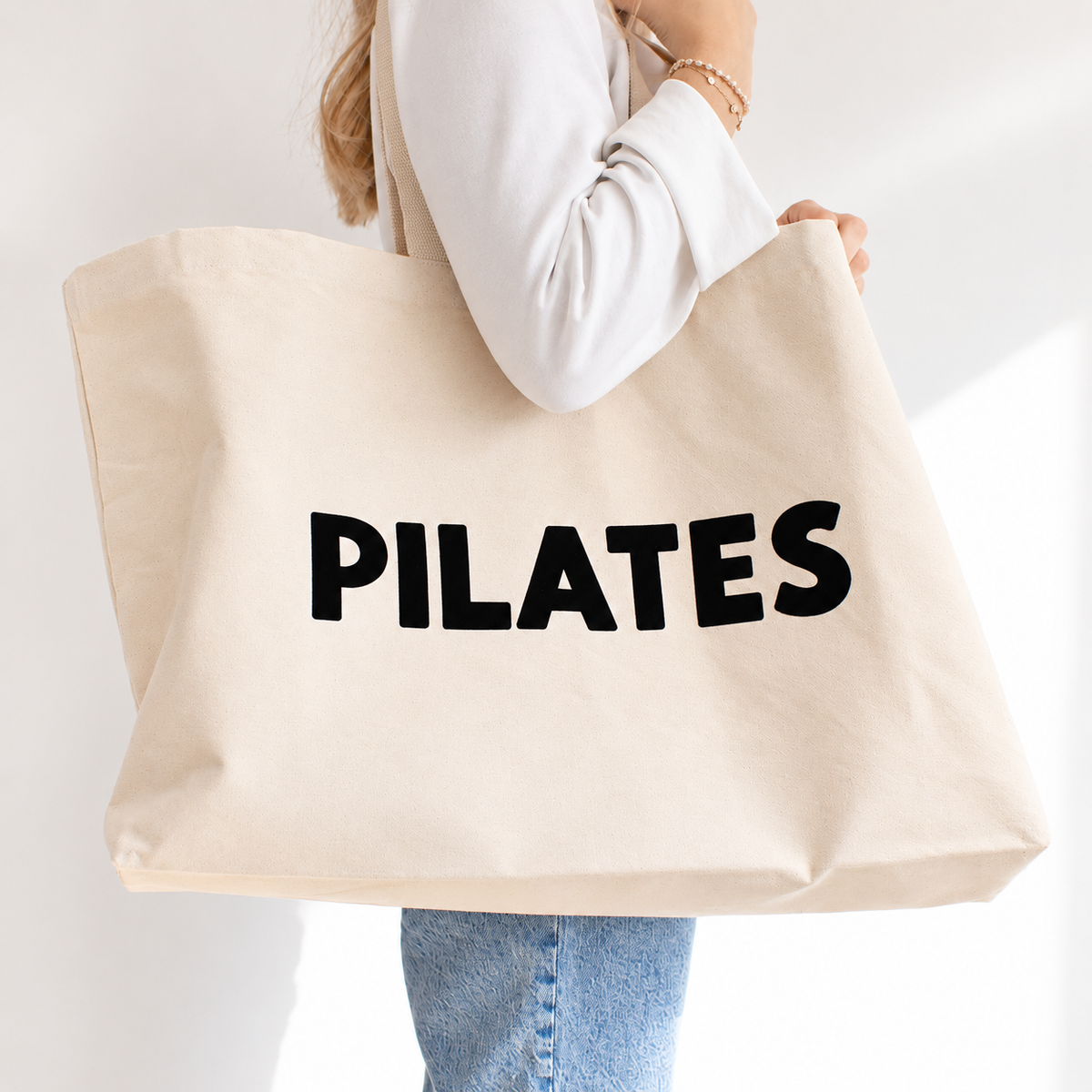 A person holding a large natural beige canvas tote bag with the word 'PILATES' printed in bold black letters across the front, against a clean white background.
