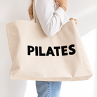 A person holding a large natural beige canvas tote bag with the word 'PILATES' printed in bold black letters across the front, against a clean white background.