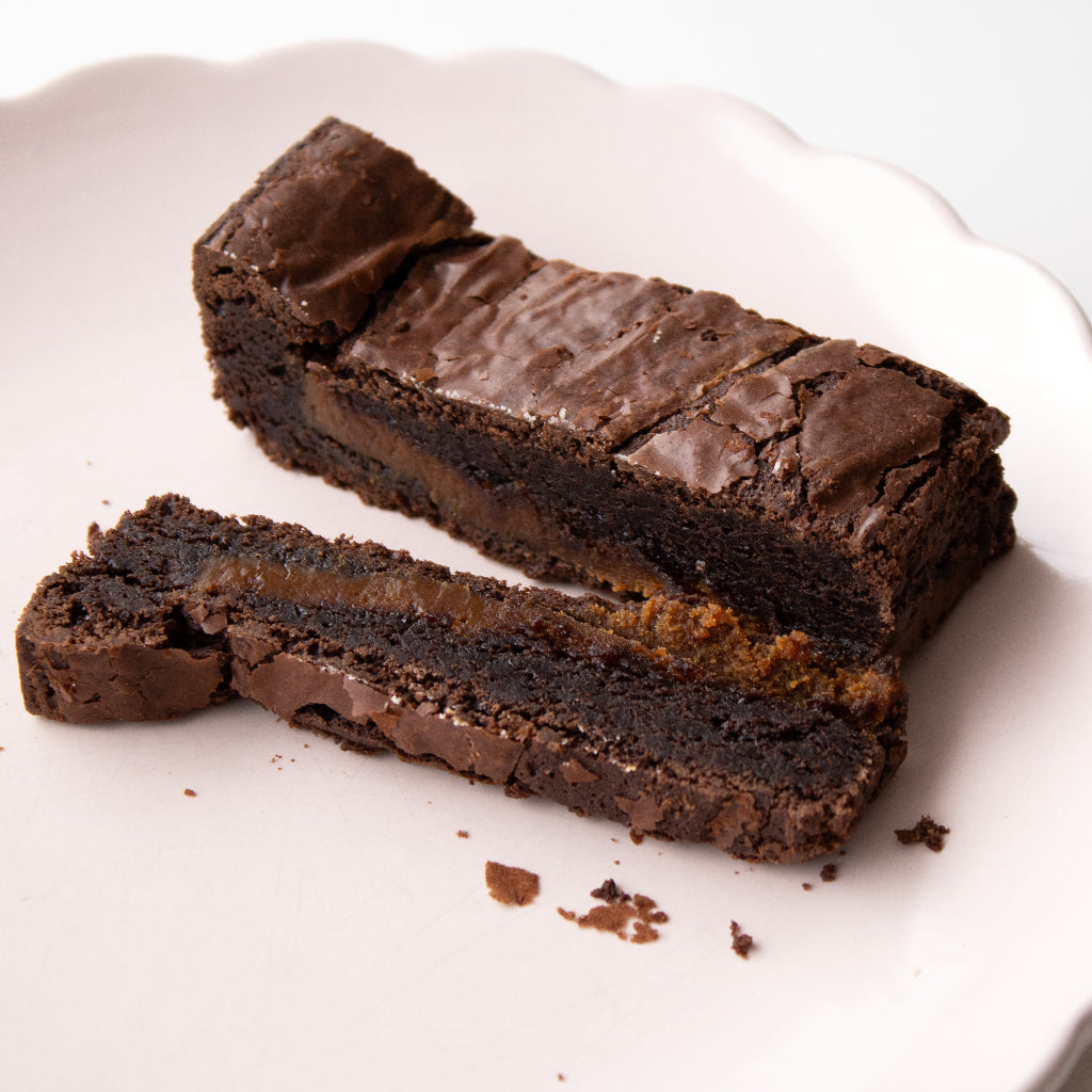 A close-up of a chocolate brownie with a thick layer of salted caramel filling, cut into pieces on a white scalloped plate.