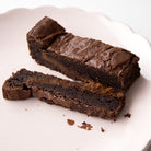 A close-up of a chocolate brownie with a thick layer of salted caramel filling, cut into pieces on a white scalloped plate.