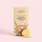 A beige box of Cartwright & Butler Sea Salt & Pepper Savoury Wafers for Cheese, shown on a solid light pink background with two wafers in front.