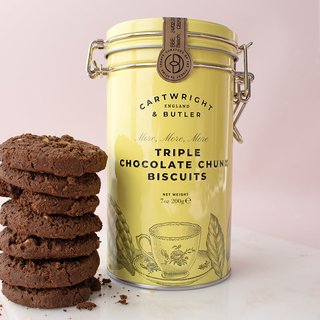Yellow canister of Cartwright & Butler Triple Chocolate Chunk Biscuits with cookies on a light pink background