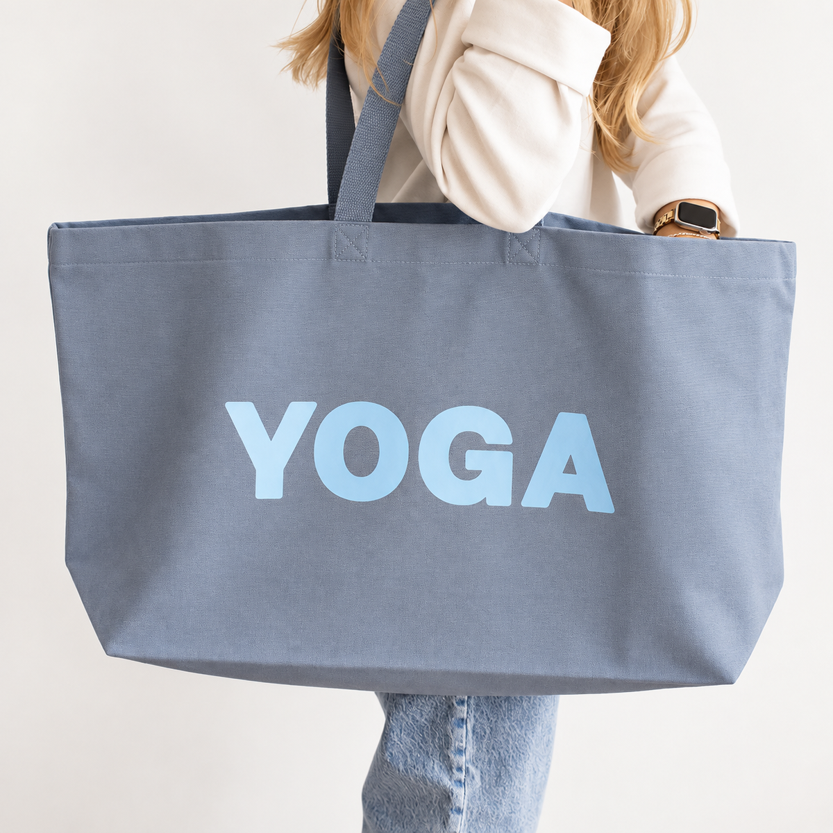 A person holding a large slate blue canvas tote bag with the word 'YOGA' printed in light blue across the front, against a plain off-white background.