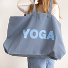 A person carrying a large slate blue canvas tote bag with the word 'YOGA' printed in light blue across the front, shown against a plain off-white background.