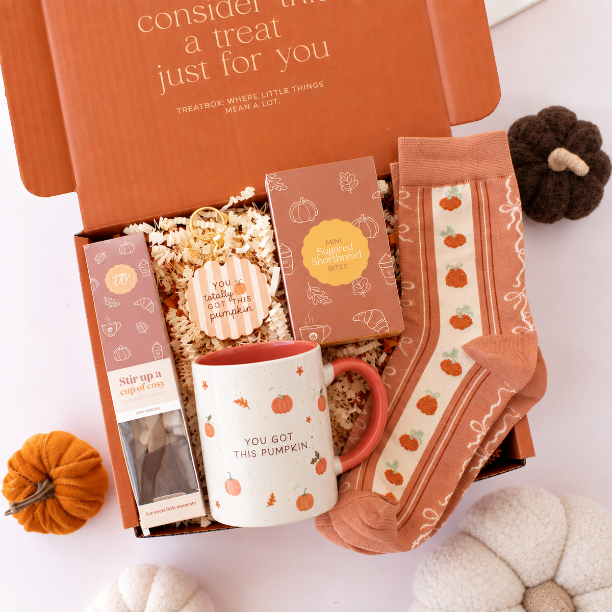 An autumn-themed gift box set from Treatbox, featuring a white ceramic mug with a pumpkin design, patterned socks, a pumpkin-shaped candle, and a tea bag, arranged in an open orange box with decorative pumpkins on a white background.